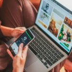Person using a laptop and smartphone simultaneously indoors, showcasing multitasking.