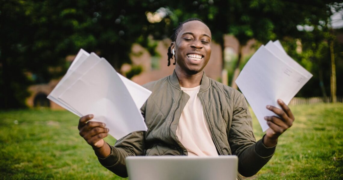 Happy young man holding papers with a laptop outdoors, showcasing academic achievement and joy.