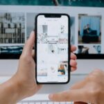 Person using smartphone to browse photos, office desktop setup.
